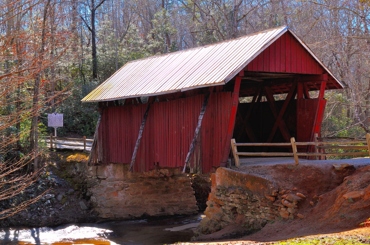 Campbell’s Covered Bridge - a red covered bridge with a gabled roof. The bridge spans across a small creek, supported by stone abutments. The surrounding area is lush with leafless deciduous trees, suggesting it might be autumn or winter. A wooden fence runs along one side of the bridge, adding to the rustic charm. To the left of the frame, there’s a signpost.