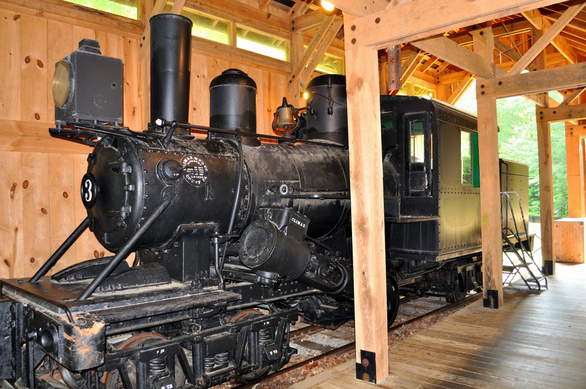 Cradle of Forestry Forest Festival Trail - old black steam locomotive with the number 3 on its side is displayed inside a wooden shelter with a green carriage attached behind it. The locomotive features a large smokestack, round headlamp, and various pipes and wheels characteristic of vintage trains. The shelter has an open design with visible wooden beams and rafters, allowing natural light to illuminate the train