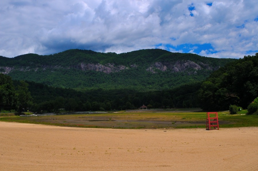 Lake Lure Beach in June 2025 - Sandy beach in front of an empty lake. A red lifeguard tower sits on the right side of the beach and mountains tower over the dry lake bed in the distance,