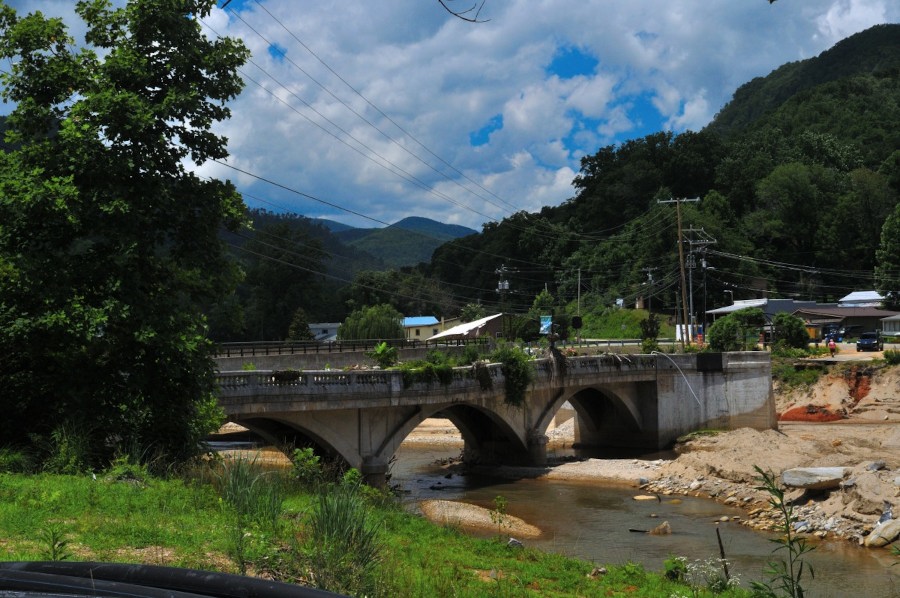 Lake Lure Flowering Bridge in June 2025 - Bridge across a muddy river with the road on one side washed away. Mountains surround the bridge/