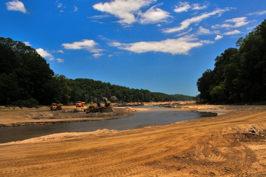Lake Lure 2025 - Dump trucks and excavators remove debris from an empty lake bed. A river snakes its way through the sandy lake bed and the entire area id flanked by rows of trees with some homes viable,