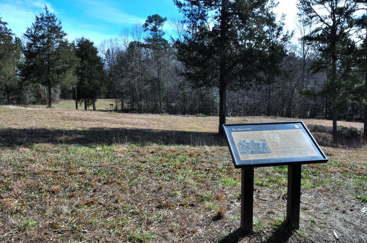 Musgrove Mill Battlefield - a serene outdoor setting with an informational sign in the foreground, surrounded by a grassy field and leafless trees, under a clear sky with scattered clouds. The text on the sign is illegible. It appears to be a historical site where visitors can learn about its significance while enjoying the natural surroundings. There’s another similar sign or structure visible in the distance to the left. The scene suggests it might be autumn or winter due to the bare branches.