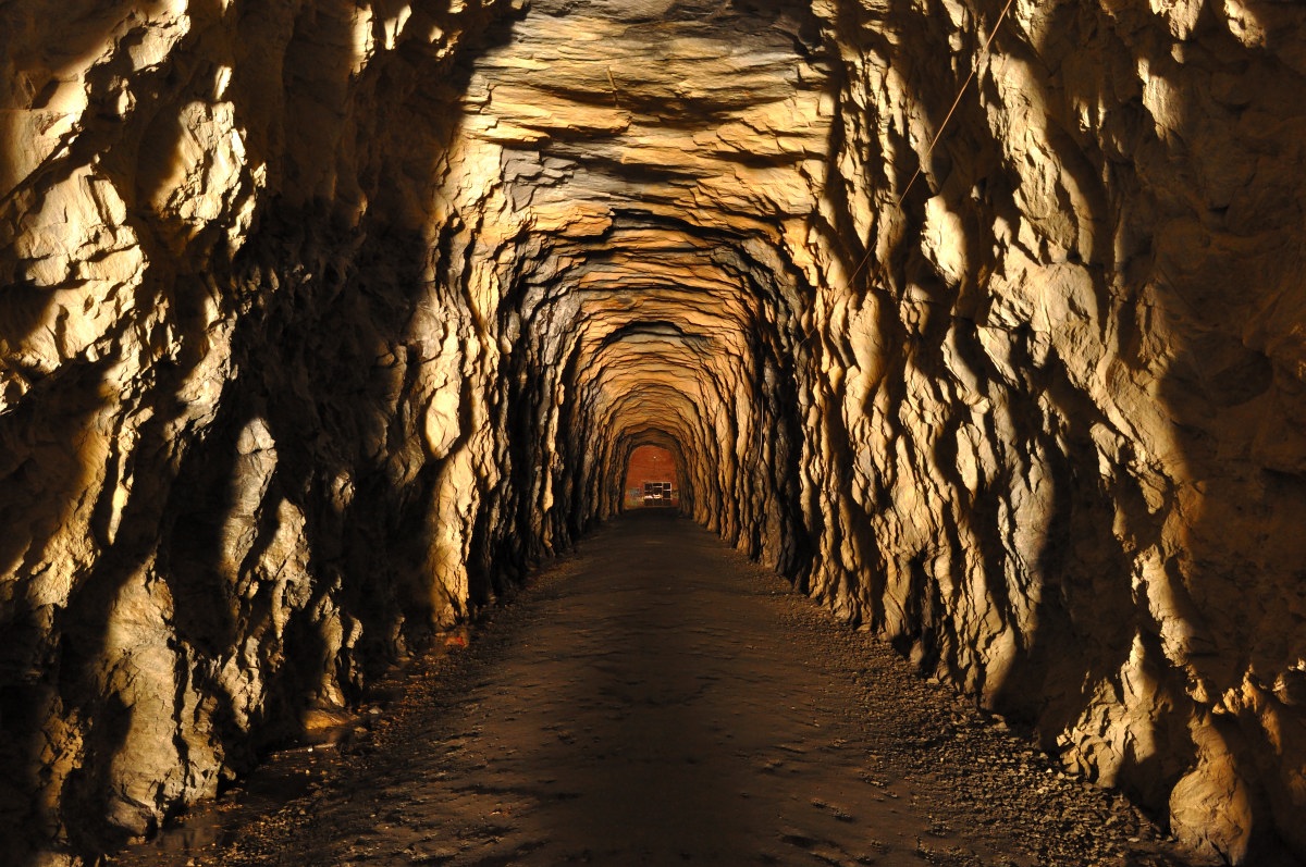 Inside the Stumphouse Mountain Tunnel - tunnel, carved through rock and illuminated by warm lighting, creates a captivating scene with its golden hues and textured walls. The image draws the viewer’s eye towards the center, where the tunnel curves slightly before reaching an opening in the distance. The natural striations and irregularities in the rock are highlighted by the interplay of light and shadow,