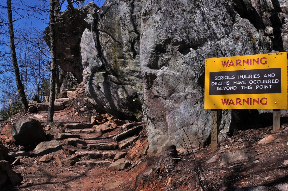A cautionary outdoor scene with a rocky, uneven terrain and a steep, narrow dirt path leading up alongside a large rock formation. Leafless trees and a clear blue sky form the backdrop. A prominent yellow warning sign with black and red text stands out, cautioning: ‘WARNING SERIOUS INJURIES AND DEATHS HAVE OCCURRED BEYOND THIS POINT.’