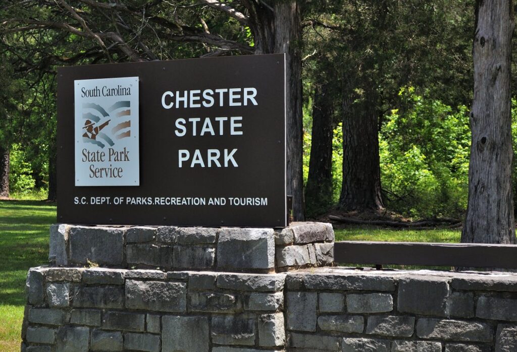 Chester State Park - sign for Chester State Park, managed by the South Carolina State Park Service. The sign is black with white lettering and features the South Carolina State Park Service logo on the top left corner, which includes an image of a palmetto tree and crescent. It is mounted on a stone structure with green trees and grass in the background under a clear sky.