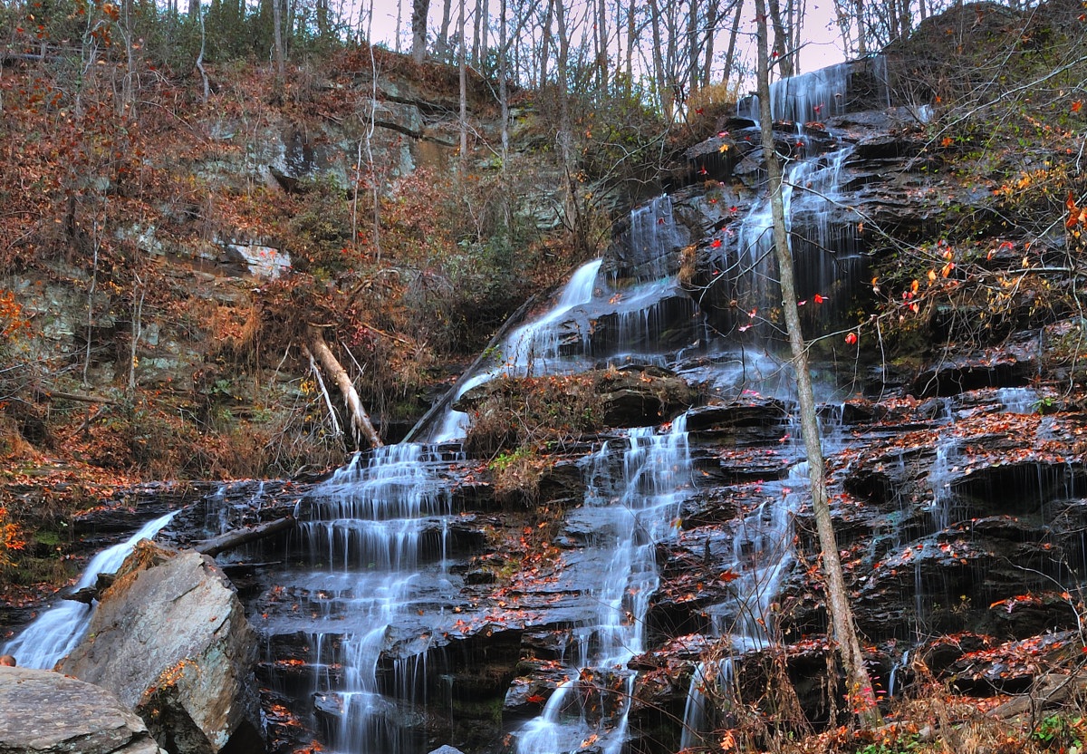 Issaqueena Falls - a cascading waterfall flowing over a rugged, multi-tiered rock face surrounded by autumn foliage. The water streams down in thin, white veils against the dark, wet stones, creating a serene and picturesque natural scene. The trees around the waterfall are sparsely dotted with orange and red leaves, indicating a late fall season
