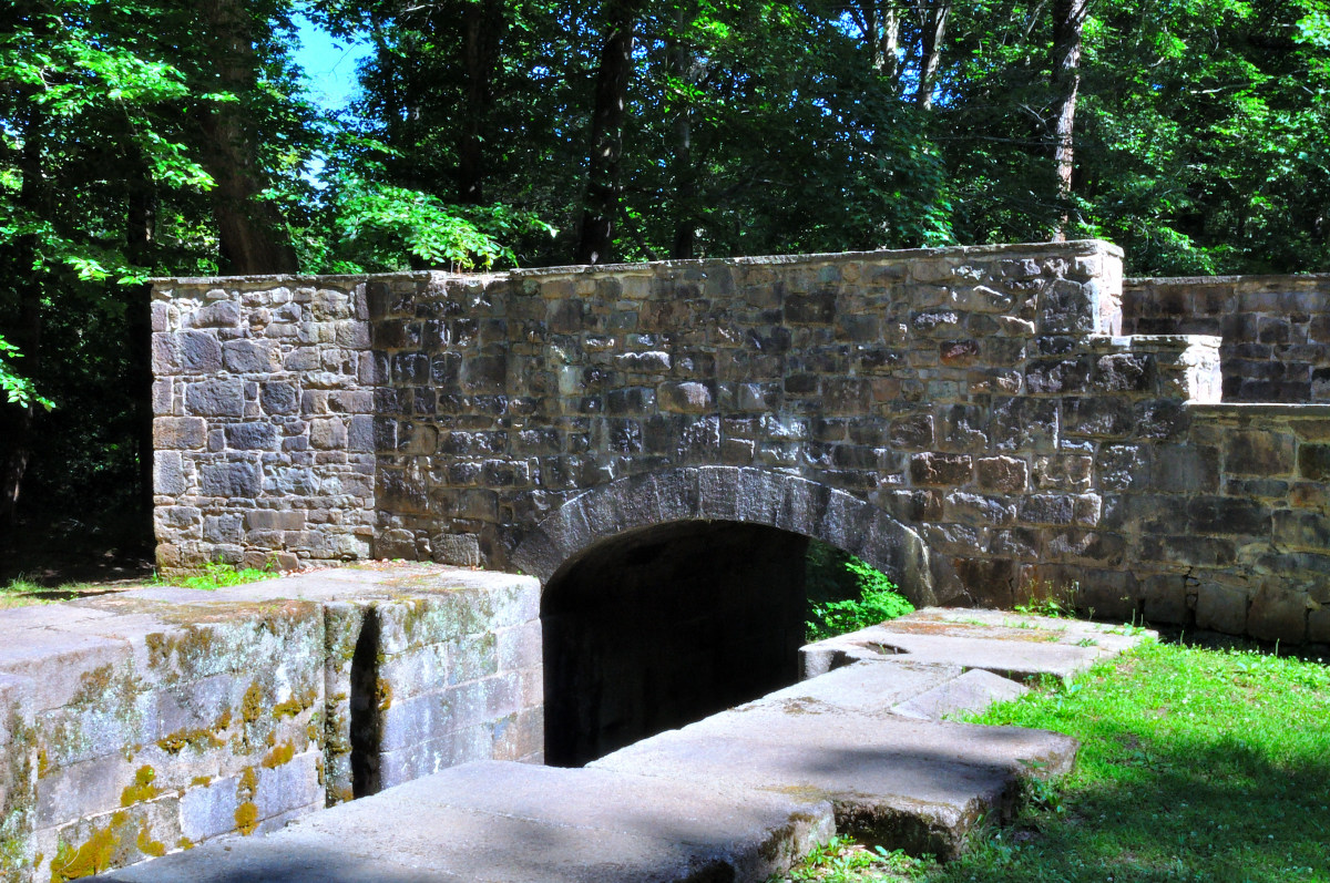 Landsford Canal - an old stone bridge over a small waterway, surrounded by lush greenery. The bridge features a single archway with moss and lichen growth on the stones, indicating age and weathering. The stonework has various shades of gray and brown, with some stones jutting out slightly more than others, giving the structure texture and character.