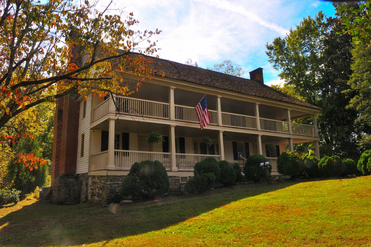 Historic Carson House Front - a two-story historic house with a large, covered front porch supported by white columns. The upper level of the porch extends across the length of the house, featuring wooden railings and visible through the gaps are multiple windows with dark shutters. An American flag hangs prominently from the center of the upper porch. The house is constructed with a combination of stone on the lower part and white siding on the upper part, topped with a dark shingled roof. Surrounding the house is a well-kept lawn with scattered trees showing early autumn colors