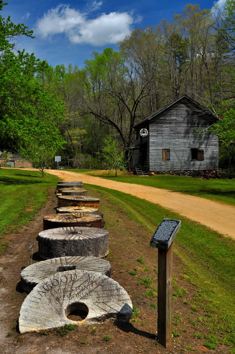 At Hagood Mill Historic Site, a line of large, engraved millstones rests on a grassy lawn, the nearest marked “WOODMAN Mill Works.” A small interpretive plaque stands beside them. Behind the stones, a weathered wooden mill building with gray siding and a circular emblem faces a winding dirt path, framed by leafy green trees and a partly cloudy blue sky