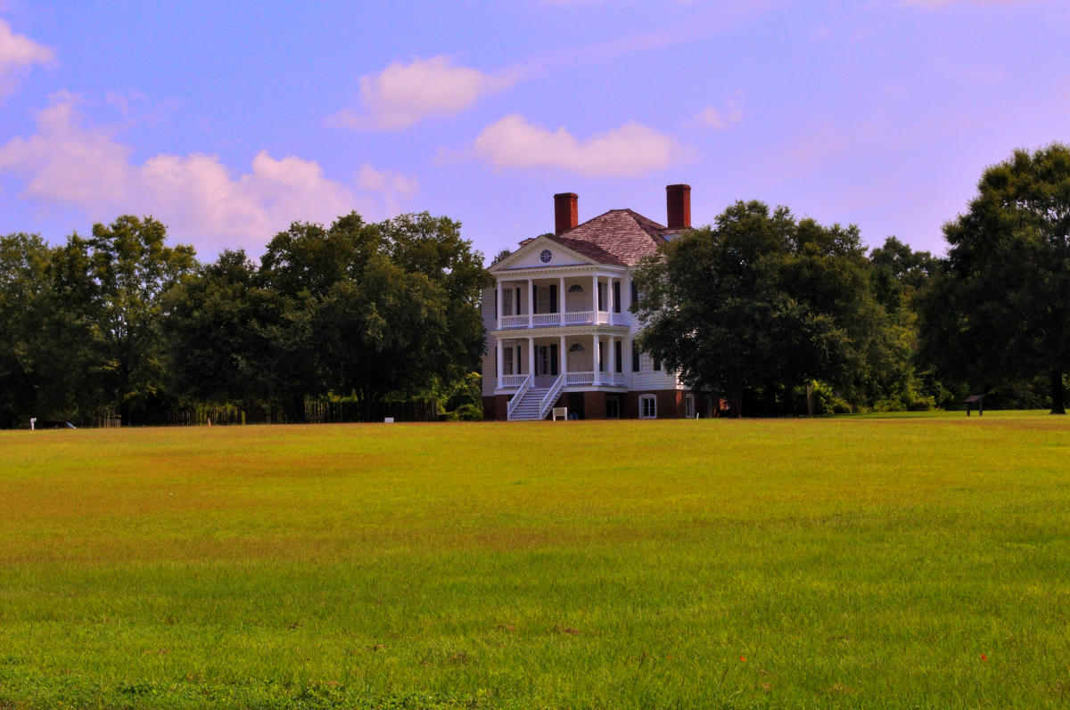 The Kershaw-Cornwallis House at the Camden Revolutionary War Site in South Carolina. A large white two-story home with tall columns, twin brick chimneys, and a central staircase, surrounded by tall trees and open grassy fields beneath a partly cloudy sky.