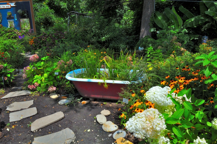 A whimsical garden display centered around a vintage red clawfoot bathtub repurposed as a water feature, overflowing with aquatic plants. Surrounding the tub are vibrant orange coneflowers, clusters of white hydrangeas, and a mix of lush greenery. A stone path winds through the scene, inviting viewers into the densely planted space. In the background, a playful blue-painted wall with a small window and shelf adds a cozy, artistic flair to the garden’s layout.
