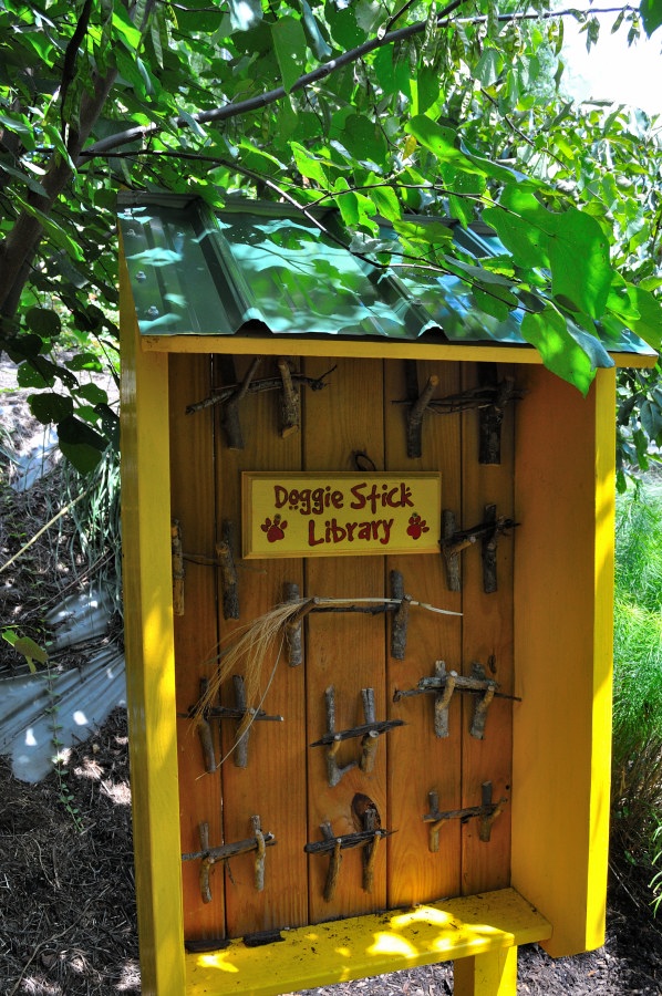 A charming outdoor installation labeled "Doggie Stick Library" stands amid leafy green plants. The small yellow wooden structure has a green metal roof and features neatly arranged wooden sticks hanging on its front in a grid-like pattern. Above the sticks, a playful red sign with paw prints reads “Doggie Stick Library,” inviting canine visitors to take a stick and enjoy. The surrounding garden space creates a whimsical and welcoming environment for pets and nature lovers alike.