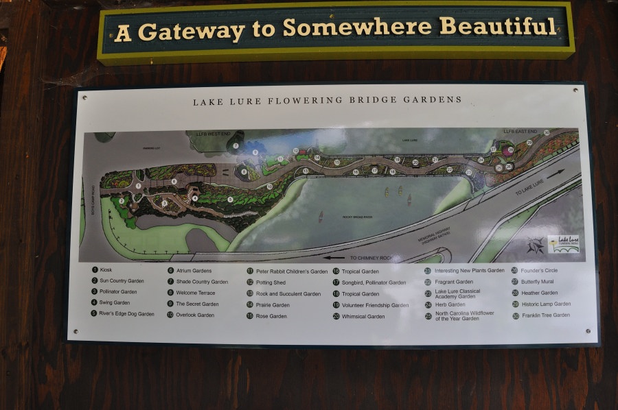 A decorative signboard showcasing a vibrant illustrated map of the Lake Lure Flowering Bridge Gardens. At the top, a green-and-yellow banner reads “A Gateway to Somewhere Beautiful,” evoking a warm and inviting atmosphere. Below, the detailed map outlines the bridge’s garden layout, featuring labeled sections such as the Sun Country Garden, Butterfly Mural, Peter Rabbit Children's Garden, and Historic Lamp Garden, among many others. A legend identifies over two dozen themed garden areas, connected by paths and nestled near Lake Lure’s scenic waters and nearby roads.