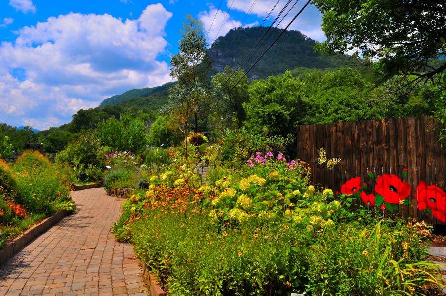A lush garden path meanders through a riot of vibrant spring blooms and greenery. On the right, a rustic wooden fence is painted with cheerful red poppies and a bright yellow butterfly, lending a whimsical, handcrafted touch to the scene. Yellow and orange flowers flourish in planter beds and hanging baskets along the path, while distant rolling hills and a partly cloudy sky provide a tranquil natural backdrop. Overhead, utility lines stretch across the horizon, subtly blending modern elements with the garden’s serene charm.