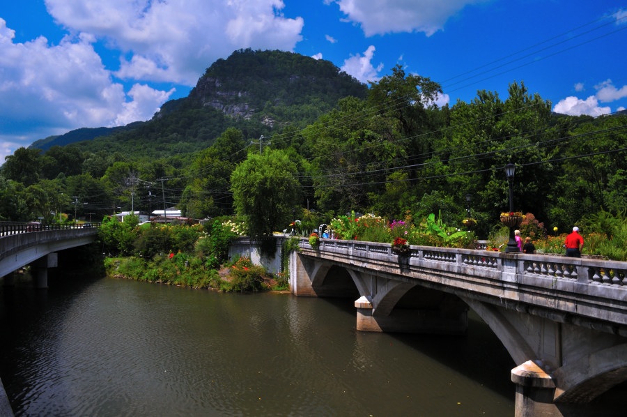 Two bridges side by side over a river. One Bridge has plants and flowers and people walking over while the other has traffic. A large mountain rises above the pair in the distance.