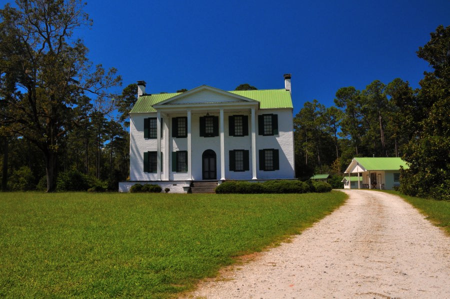 Two-story white Federal Style home with a green metal roof, black shutters, and a columned portico at Belfast Wildlife Management Area, surrounded by trees and open lawn.