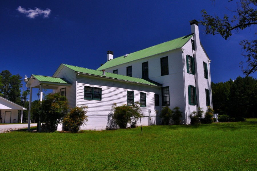 Large white house with a green metal roof and green shutters at Belfast Wildlife Management Area, surrounded by grass and trees under a clear blue sky.