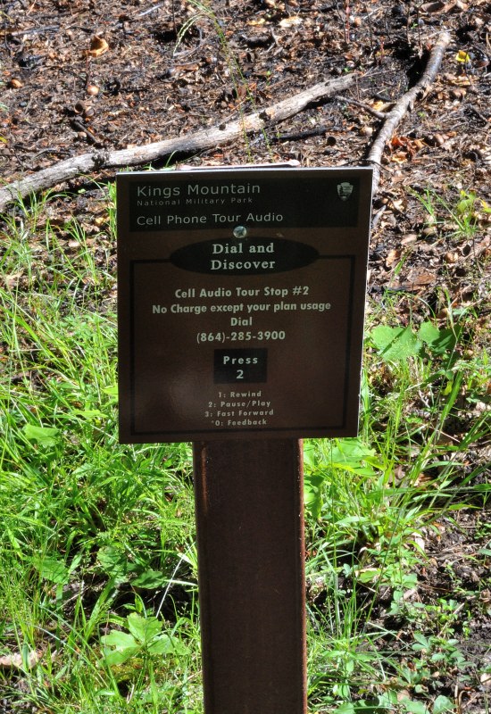 Informational sign at Kings Mountain National Military Park offering a cell phone audio tour, with instructions for dialing and accessing stop #2, set against a natural forest backdrop.