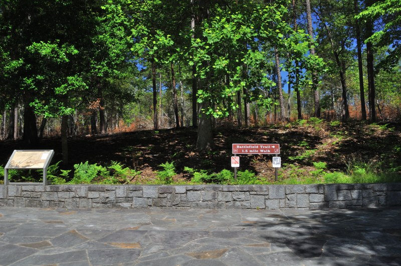 Trailhead sign at Kings Mountain National Military Park marking the Battlefield Trail, a 1.6‑mile walk with restrictions against bicycles and pets, set beside an informational display and surrounded by forest greenery