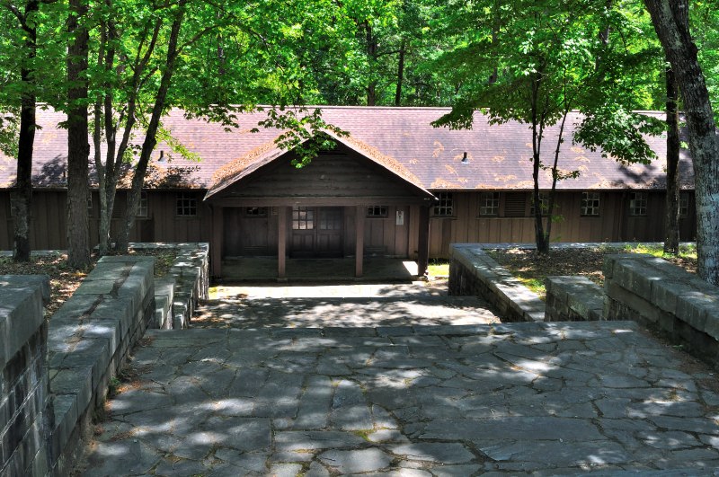 The bathhouse at Kings Mountain State Park stands nestled among tall trees, its rustic wooden structure featuring a gabled shingle roof and a covered entrance supported by timber beams. Stone walls flank the building, and a wide stone path leads to the doorway. Surrounded by forest and dappled sunlight, the bathhouse blends traditional craftsmanship with the serenity of its natural setting.