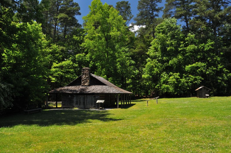 A rustic log blacksmith shed stands in a sunlit clearing at Kings Mountain State Park. The structure features a stone chimney, wooden walls, and a simple porch supported by posts. A small interpretive sign sits out front, while another wooden outbuilding is visible in the background. Tall trees surround the clearing, framing the shed as part of the park’s living history farm and evoking the craftsmanship of Carolina’s past.